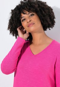 Maglietta lunga a maniche con scollo a V di colore rosa, realizzata in un tessuto morbido con una leggera texture. Le maniche arrivano ai polsi, con una vestibilità casual e rilassata.