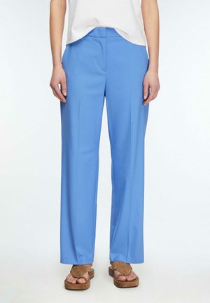 Kobieta ubrana w jasnoniebieskie spodnie o szerokich nogawkach, biały top i brązowe sandały na platformie, stojąca na białym tle.