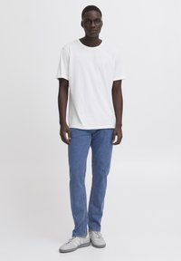 T-shirt bianca in cotone con scollatura rotonda, abbinata a jeans blu in denim e sneakers grigie, in piedi contro uno sfondo neutro.