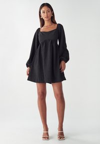 Schwarzes Kleid mit quadratischem Ausschnitt, langen Puffärmeln und schwingendem Rock. Strukturierter Stoff mit subtilen Musterdetails. Weiße Riemchen-Highheels.