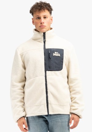 Crème fleecejack met een marineblauwe voorzak. Hoge kraag, ritssluiting en zachte textuur. Bevat een logo op het zakgebied.