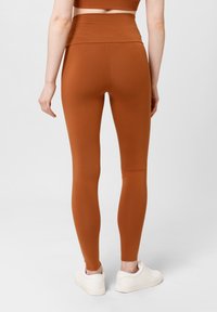 Leggings marrones de talle alto hechos de una tela elástica, diseño ajustado, textura suave y construcción sin costuras, combinados con zapatillas blancas.