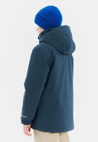 Marineblauwe waterdichte jas met een capuchon, elastische manchetten en een klein logo op de mouw. Draag met een heldere blauwe gebreide beanie.