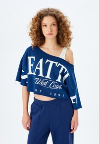 Blå kort sweatshirt med vit text "SEATTLE West Coast EST. 1991", lös passform, off-shoulder design och rå kant. Matchad med marinblå byxor.