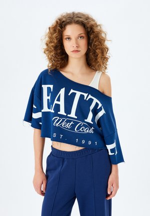 Blauwe cropped sweatshirt met witte opdruk "SEATTLE West Coast EST. 1991", losse pasvorm, off-shoulder ontwerp en raw-edgezoom. Gecombineerd met navy broeken.