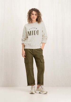 Lichtgrijze sweatshirt met "APRES MIDI" opdruk, olijfgroene cargobroek met zijzakken, en beige sneakers. Model poseert tegen een neutrale achtergrond.