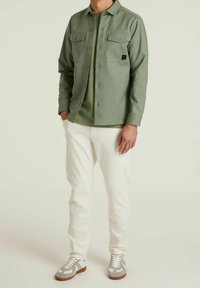 Lichtgroen overhemd met knopen en twee borstzakken, gedragen over een bijpassend groen shirt, gecombineerd met witte broeken en grijze sneakers.