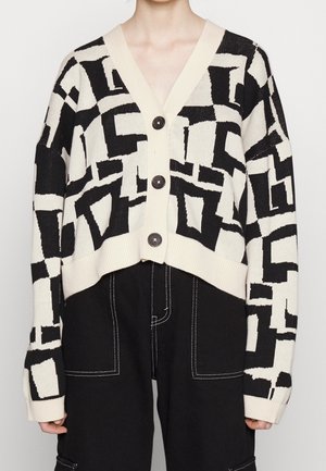 Schwarz-weiß gemusterte, geometrisch gemusterte, geknöpfte Strickjacke, getragen über schwarzen Hosen mit weißen Nähten.