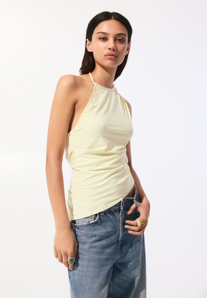 Top sans manches jaune clair avec un design à col halter. Texture lisse, côtés drapés, associé à un jean en denim taille haute.