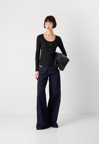 Zwarte langemouw top met een ronde hals, gecombineerd met donkerblauwe wijde denim jeans. Model houdt een zwarte leren tas vast.