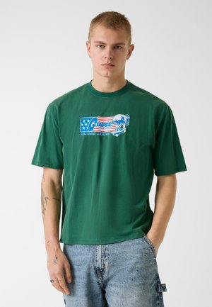 Junger Mann mit kurzen blonden Haaren, der ein grünes T-Shirt mit rotem, weißem und blauem Motiv und hellblauen Jeans trägt, vor einem schlichten Hintergrund.