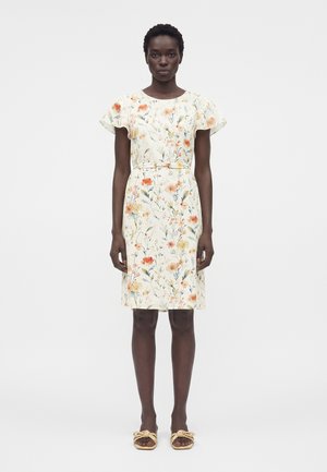 Žena v bielej šatách po kolená s oranžovo-žltými kvetinovými vzormi, rozstrapkanými rukávmi a béžovými slip-on sandálmi s detailmi z lana.