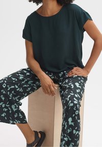 Haut vert foncé à manches courtes assorti à un pantalon large à motifs noirs et bleu clair. Modèle assis sur un socle en bois, portant des sandales noires.