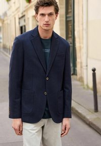 Blazer en laine bleu marine avec deux poches avant et une fermeture à un bouton, porté sur un t-shirt vert foncé et un pantalon gris clair.