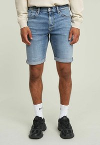 Pantalones cortos de mezclilla azul claro con dobladillo enrollado, cinco bolsillos y un clásico diseño de cremallera. Combinados con zapatillas negras y calcetines blancos.