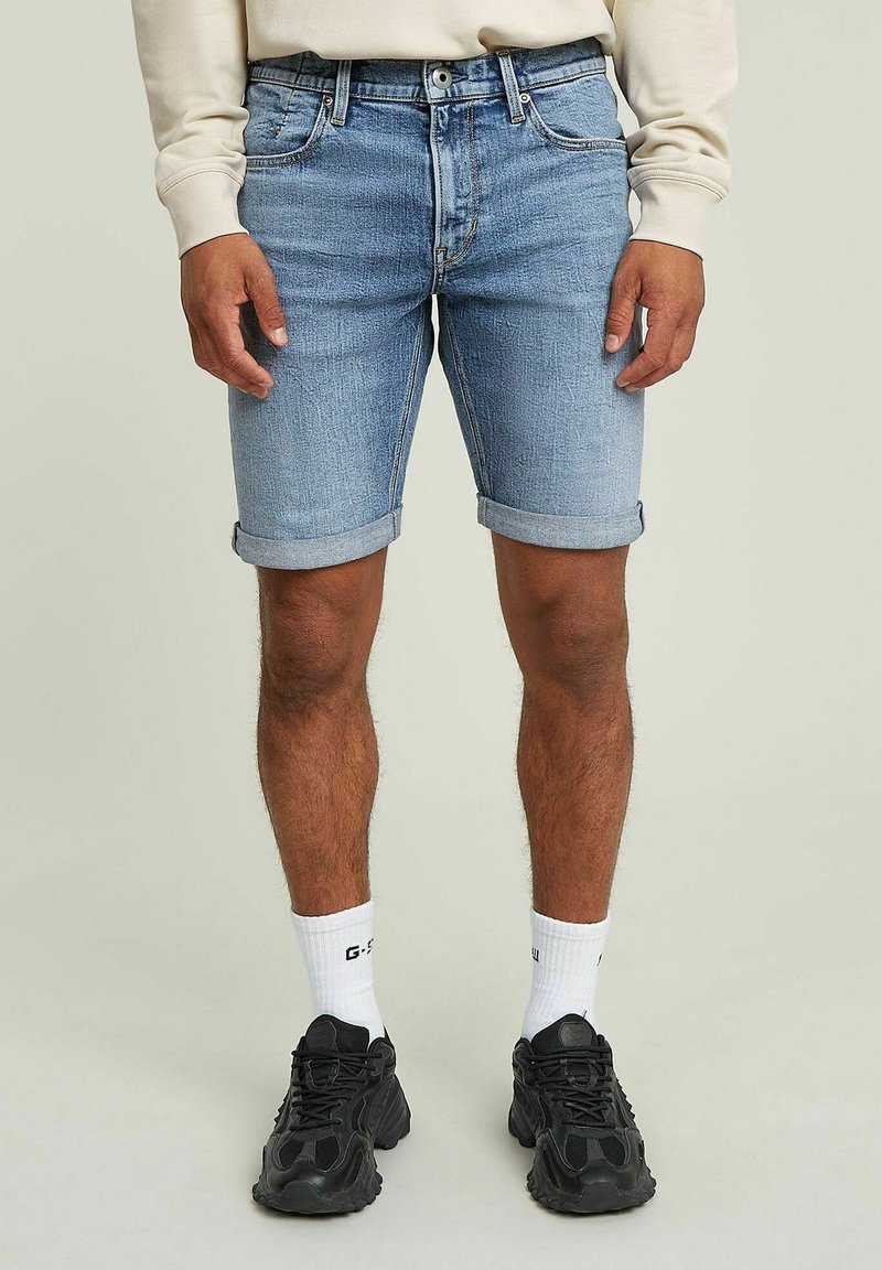 Pantalones cortos de mezclilla azul claro con dobladillo enrollado, cinco bolsillos y un clásico diseño de cremallera. Combinados con zapatillas negras y calcetines blancos.
