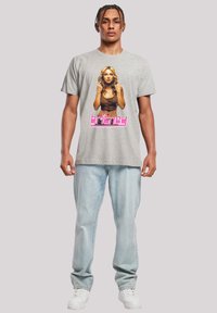 Graues kurzärmliges T-Shirt mit einem Grafikdruck einer Frau in einem braunen Oberteil, darunter in rosa der Text "Britney". Kombiniert mit hellblauen Jeans und weißen Sneakers.