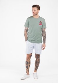 Mężczyzna z pełnymi tatuażami na ramionach i nogach, ubrany w zielony t-shirt z nadrukiem palm, w białych szortach, stojący z dłońmi w kieszeniach, patrzący w lewo.