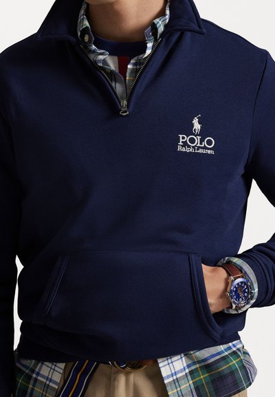 Pulôver de polar azul-marinho com colarinho de fecho de correr de um quarto, com um bolso frontal e um logótipo branco bordado. Camisa xadrez por baixo, relógio casual visível.