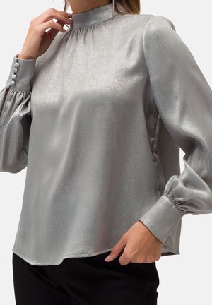 Mujer con blusa plateada brillante de manga larga, cuello alto y puños abotonados, combinada con pantalones negros, posando contra un fondo liso.