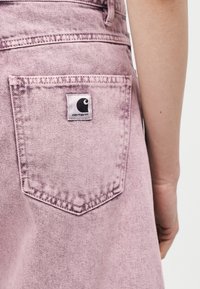 Κοντινό πλάνο ενός ατόμου που φοράει τζιν παντελόνι σε ανοιχτό μωβ χρώμα με πίσω τσέπη που φέρει ετικέτα με το λογότυπο Carhartt.