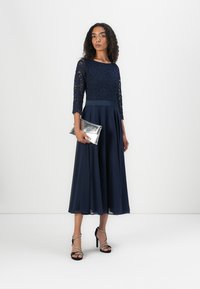 Robe en dentelle bleu marine à manches trois quarts et jupe fluide. Assortie à une pochette argentée et des escarpins noirs à lanières.