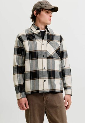 Jeune homme portant une chemise à carreaux grise et noire boutonnée, un pantalon marron et une casquette sombre, regardant vers sa gauche.