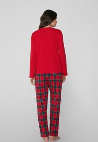 Rode top met lange mouwen gecombineerd met rood-groen geruite pyjamabroek. Zachte stof, losse pasvorm en elastische tailleband aan de broek.