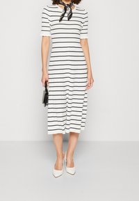 Frau in einem weißen Midikleid mit schwarzen horizontalen Streifen, weißen Absätzen, einem schwarzen Halstuch und einer kleinen schwarzen Handtasche.