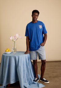 Blauwe katoenen t-shirt met 'CIAO' grafiek, gecombineerd met lichte denim shorts. Model staat naast een tafel met een blauwe doek, vaas en fruit.