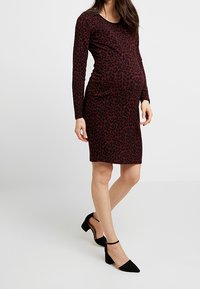 Donna incinta che indossa un abito aderente a maniche lunghe con stampa leopardata in rosso scuro e nero, abbinato a décolleté nere con cinturino alla caviglia.