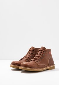 Bottines en cuir marron avec bouts ronds, fermeture à lacets, couture contrastante et semelles en caoutchouc pour une meilleure traction. Étiquette de marque rouge visible.