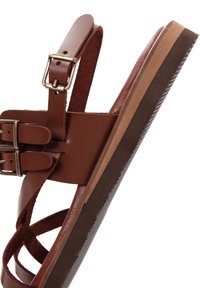 Sandale en cuir marron avec semelle plate, boucles ajustables et brides entrecroisées. Semelle intérieure texturée et semelle foncée contrastante.