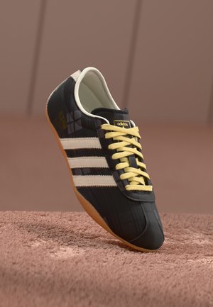Adidași negri cu șireturi galbene și dungi albe. Prezintă un material texturat și o talpă din cauciuc maro. Accentuati cu un logo pe limba.