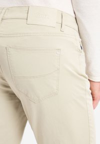 Beige katoenen broeken hebben een soepele textuur, een achterzak met een gestikte rand en een leren merkpatch op de tailleband.