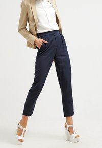 Marinblå skräddarsydda byxor med framfickor, hög midja och sidofickor, tillsammans med en vit blus och en beige kavaj, kompletterat med vita plattformsklackar.