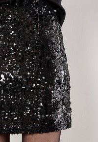 Schwarzer Pailletten-Minirock mit strukturierter Oberfläche und unterschiedlich großen Pailletten, die einen reflektierenden, schimmernden Effekt erzeugen. Glatter Bund.