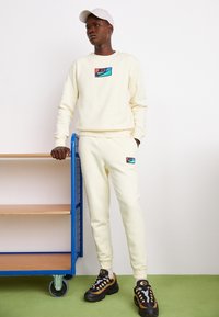 Creme sweatshirt og matchende joggers med Nike-logo; sorte og beige sneakers; står ved siden af en blå vogn mod en hvid væg.