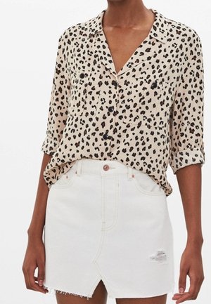 Kobieta ubrana w beżową koszulę w panterkę z podwiniętymi rękawami, wpuszczoną w białą, postrzępioną dżinsową spódnicę z przednim rozcięciem.