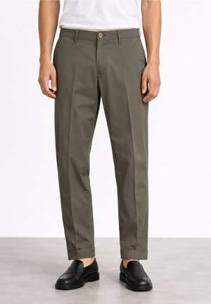 Mężczyzna w oliwkowozielonych, dopasowanych spodniach z mankietami, czarnych skórzanych mokasynach i białej, włożonej do środka koszuli, stojący na jasnoszarej podłodze.