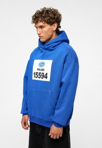 Blauwe hoodie van katoen met een voorzak, voorzien van een witte rechthoekige opdruk met "POLIZEI 15594" in zwarte letters.