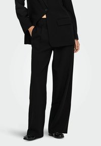 Veste de costume noire taillée avec un seul bouton, associée à un pantalon noir à jambes larges et des chaussures noires brillantes avec un accent en nœud.