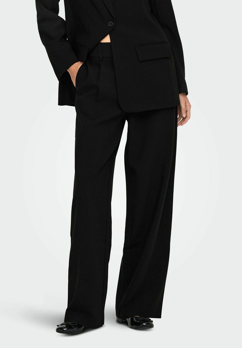 Veste de costume noire taillée avec un seul bouton, associée à un pantalon noir à jambes larges et des chaussures noires brillantes avec un accent en nœud.