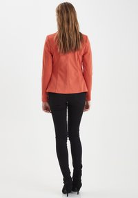 Oranžová obtiahnutá bunda s dlhými rukávmi, so švovými detailmi, nosená cez čierne úzke nohavice a členkové topánky so jemnou textúrou. Minimalistický dizajn.