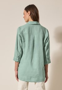 Camicia di lino verde chiaro con colletto, maniche lunghe arrotolate, dettaglio piega sul retro e vestibilità rilassata.