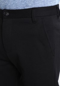 Pantalon noir avec une texture de tissu lisse, doté d'une fermeture à un bouton, de passants pour ceinture et de poches avant inclinées.