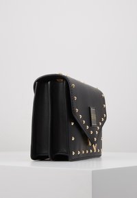 Borsa in pelle nera con borchie dorate e manico a catena, chiusura a pattina rettangolare, posizionata su un piedistallo bianco contro uno sfondo semplice.
