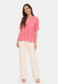 Rožinė nėrinių peplumo palaidinė su trumpomis rankovėmis ir V formos iškirpte, derinama su smėlio spalvos plačiomis kelnėmis ir bežė sandalu. Lygus paviršius ir gėlių raštas.