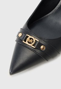Scarpa nera in pelle con punta affusolata, caratterizzata da un elemento decorativo con logo in tonalità dorata, due bande decorative e una texture liscia all'interno.