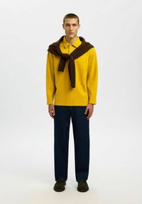 Gelbes Langarm-Polo mit strukturiertem Muster, kombiniert mit dunkelblauen weit geschnittenen Hosen. Brauner Pullover um die Schultern gebunden. Braune Schuhe.